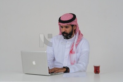 رجل سعودي يجلس ويعمل على جهاز كمبيوتر محمول ، فنجان شاي ، دراسة جدوى أعمال ، خلفية بيضاء ، موديل سعودي