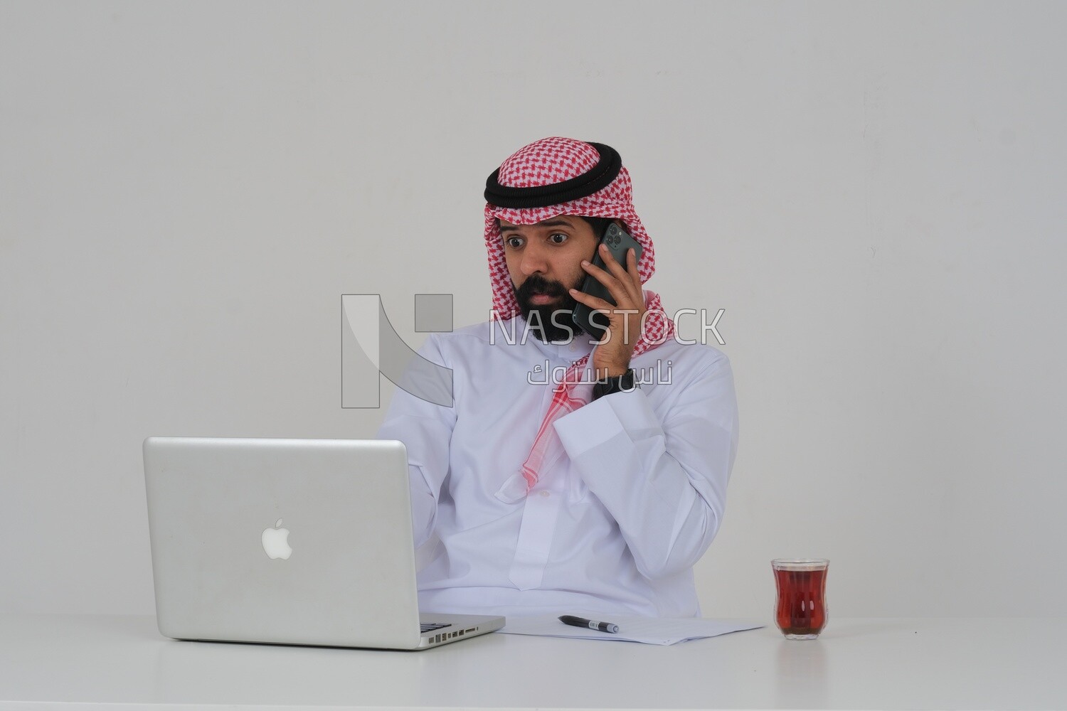 Saudi man sitting and working on a laptop, using his mobile phone, a cup of tea, business feasibility study, white background, Saudi model