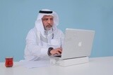 رجل سعودي يجلس ويعمل على جهاز كمبيوتر محمول ، فنجان شاي ، دراسة جدوى أعمال ، موديل سعودي