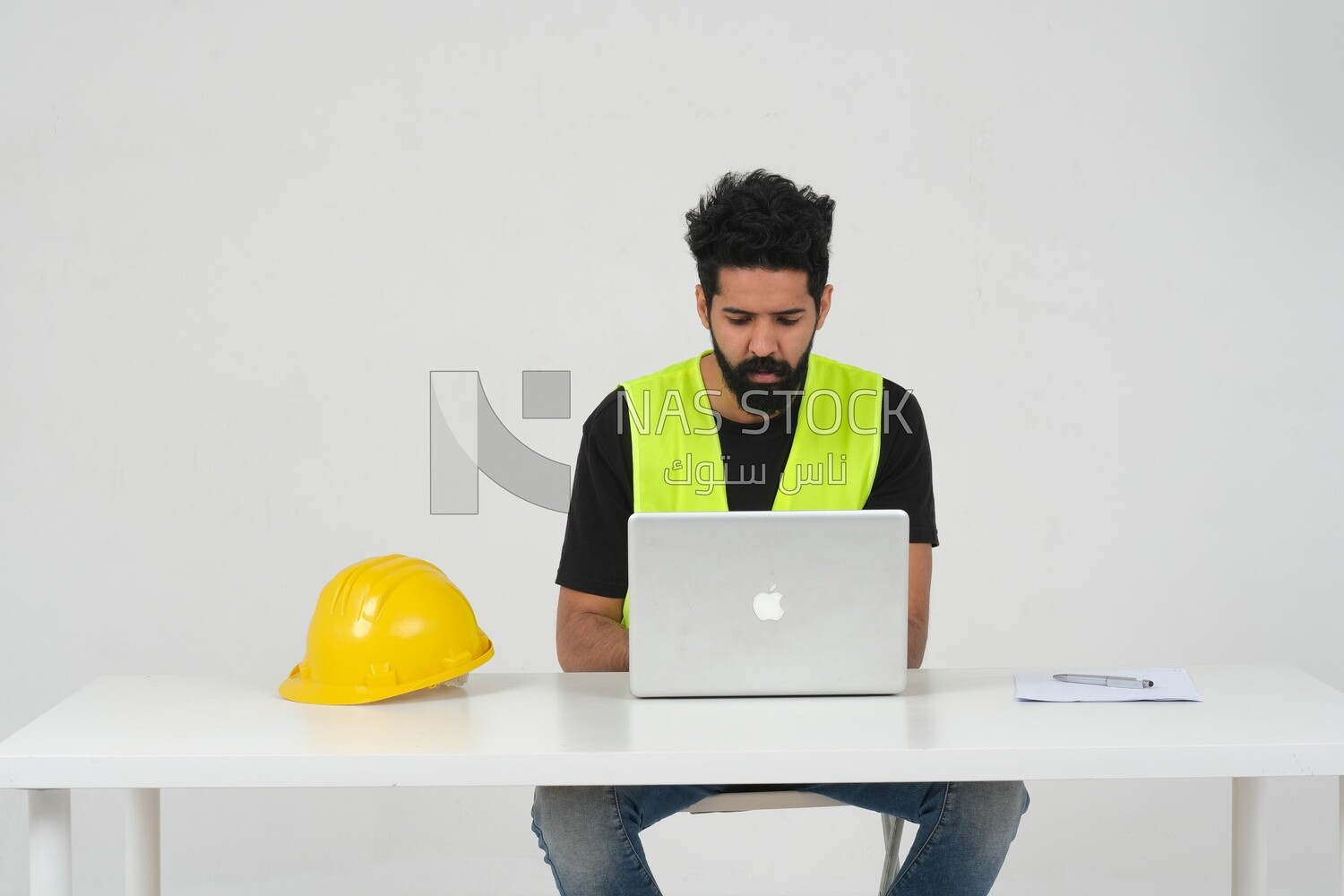 Saudi engineer wearing a safety vest sitting and working on the laptop, engineering professions and jobs, white background, Saudi model