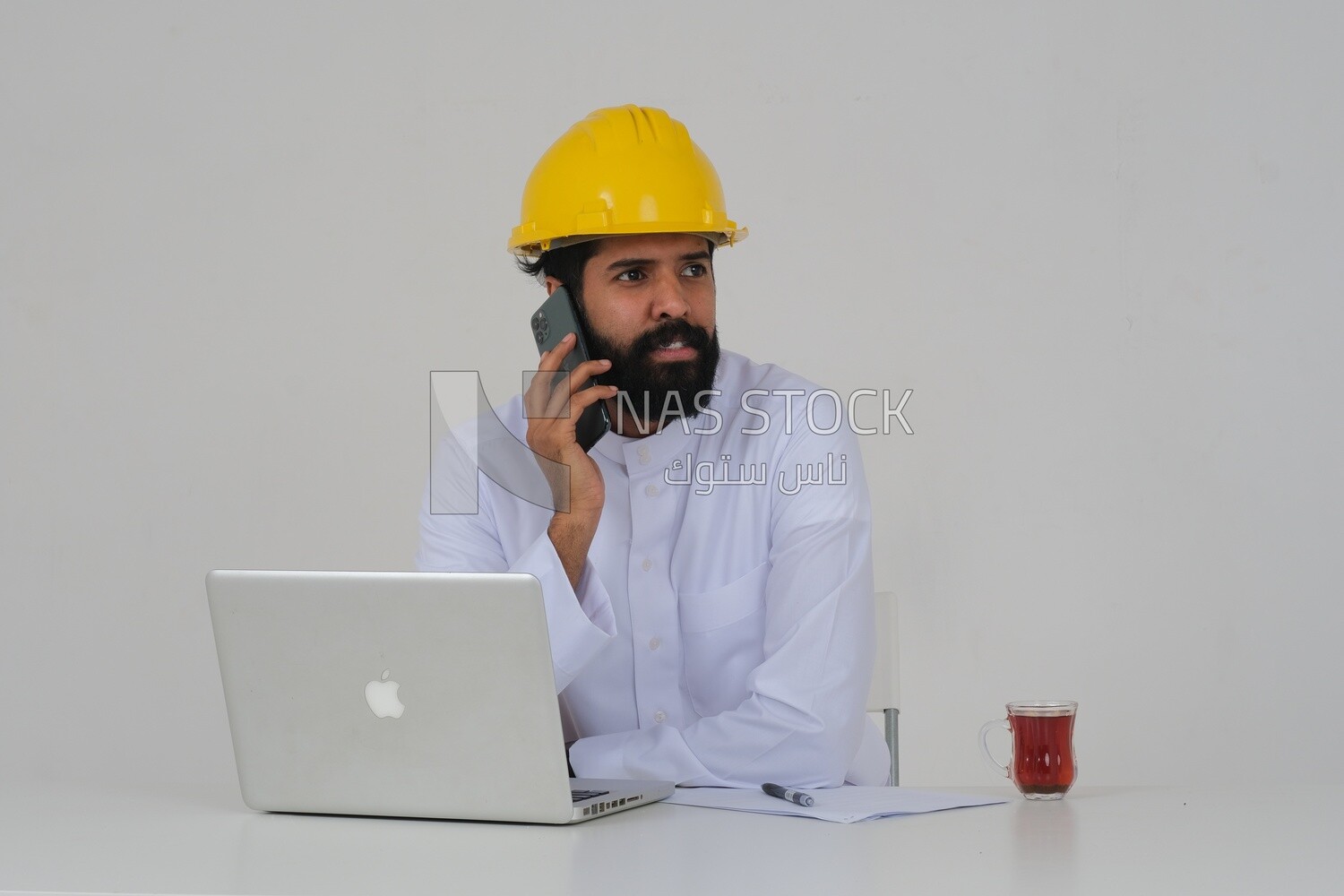 Saudi engineer wearing a work hat sitting in front of the laptop, using his mobile phone, engineering professions and jobs, white background, Saudi model