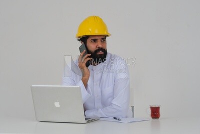 مهندس سعودي يرتدي قبعة عمل يجلس امام اللاب توب ، يستخدم هاتفه المحمول ، المهن الهندسية والوظائف ، خلفية بيضاء ، موديل سعودي