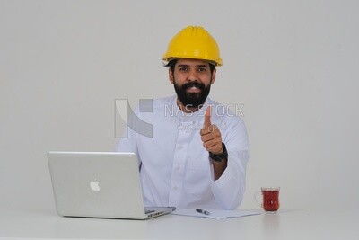 مهندس سعودي يرتدي قبعة عمل يجلس و يعمل على اللاب توب ، المهن الهندسية والوظائف ، خلفية بيضاء ، موديل سعودي