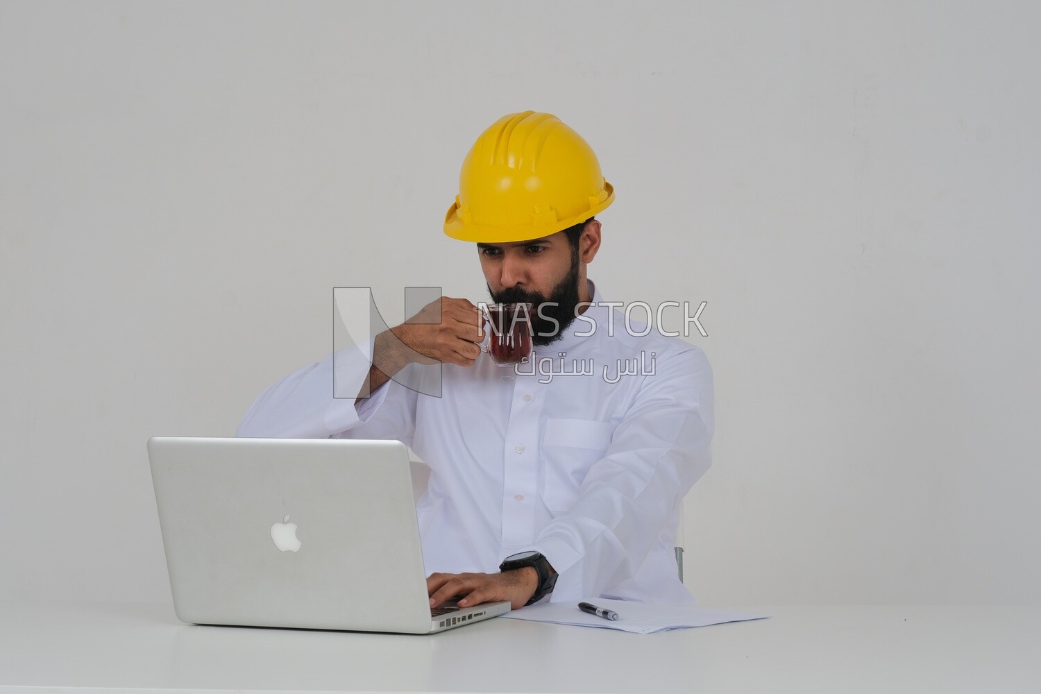 Saudi engineer wearing a work hat sitting and working on the laptop, drinking a cup of tea, engineering professions and jobs, white background, Saudi model