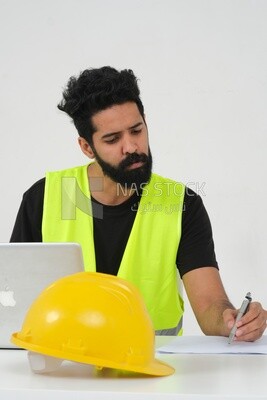Saudi engineer wearing a work hat sitting in front of the laptop, writing notes , engineering professions and jobs, white background, Saudi model