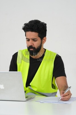 Saudi engineer wearing a work hat sitting in front of the laptop, writing notes , engineering professions and jobs, white background, Saudi model