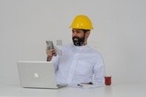 Saudi engineer wearing a work hat sitting in front of the laptop, using his mobile phone, engineering professions and jobs, white background, Saudi model