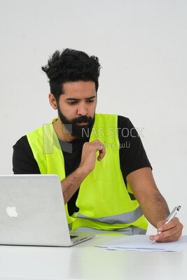 Saudi engineer wearing a work hat sitting in front of the laptop, writing notes , engineering professions and jobs, white background, Saudi model