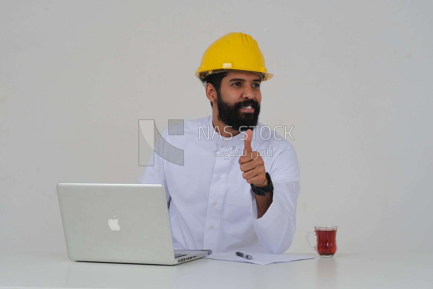 Saudi engineer wearing a work hat sitting in front of the laptop, writing notes , engineering professions and jobs, white background, Saudi model