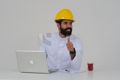 Saudi engineer wearing a work hat sitting in front of the laptop, writing notes , engineering professions and jobs, white background, Saudi model