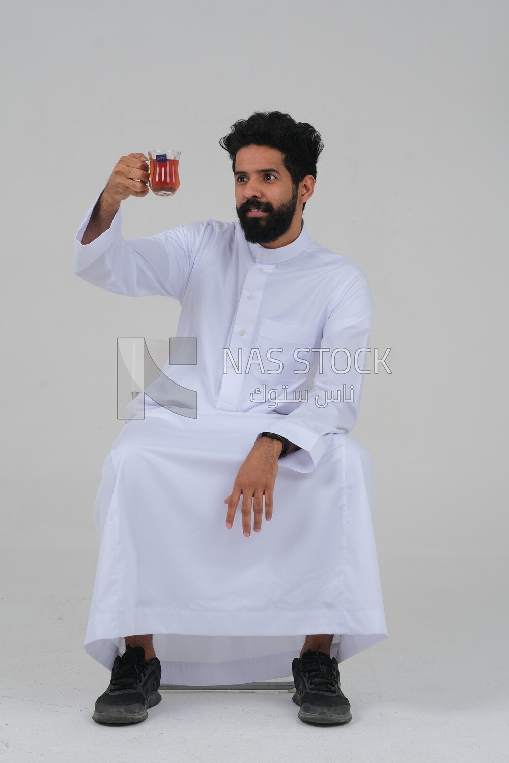 Saudi man wearing a white dress sitting on a chair, carrying a cup of tea and looking at it, self-confidence, white background, Saudi model