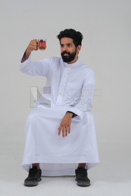 Saudi man wearing a white dress sitting on a chair, carrying a cup of tea and looking at it, self-confidence, white background, Saudi model