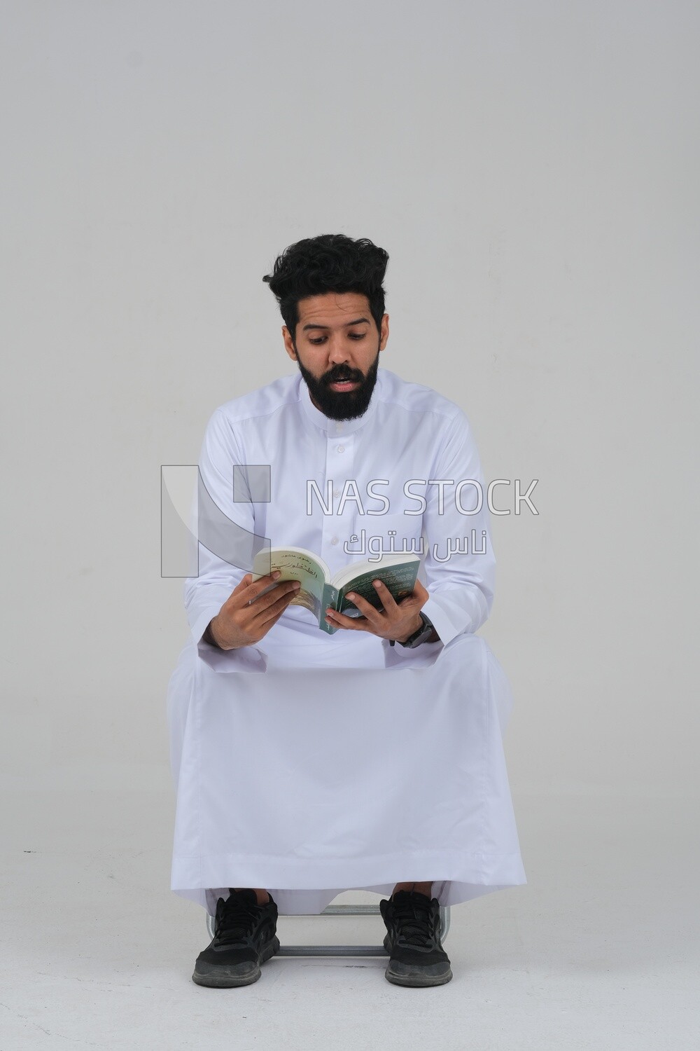 Saudi man wearing a white dress sitting on a chair, reading a book, self-confidence, white background, Saudi model