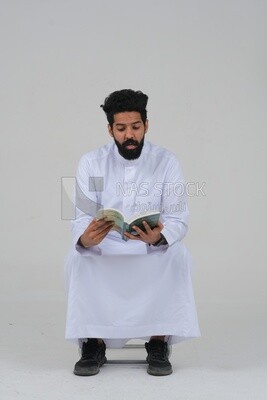 Saudi man wearing a white dress sitting on a chair, reading a book, self-confidence, white background, Saudi model
