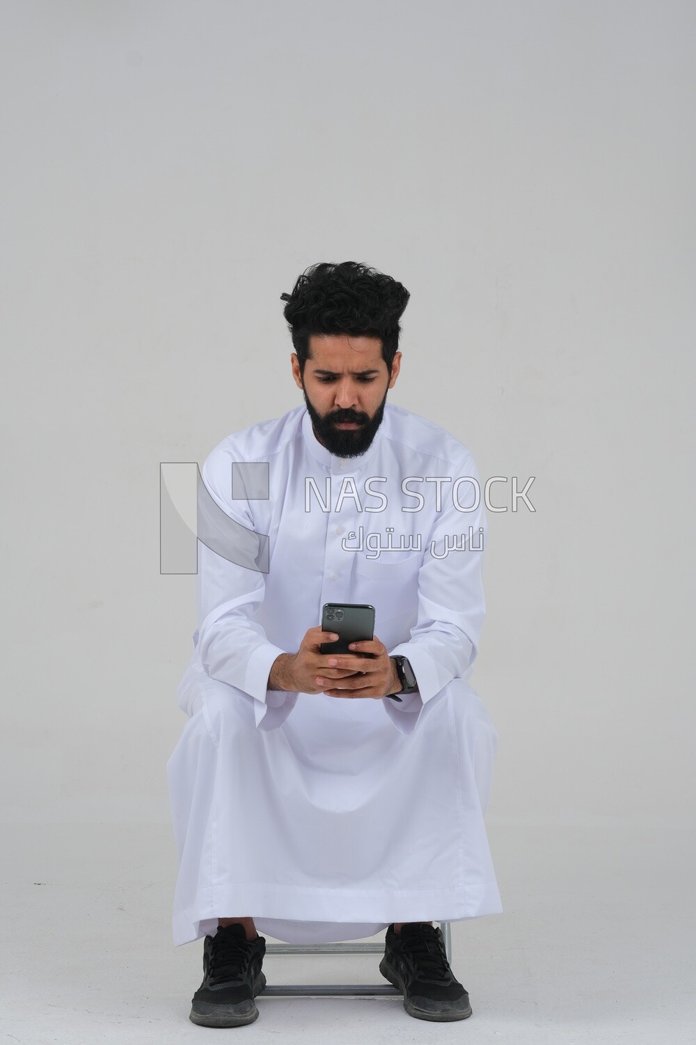 Saudi man wearing a white dress sitting on a chair, carrying his mobile phone, self-confidence, white background, Saudi model