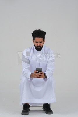 Saudi man wearing a white dress sitting on a chair, carrying his mobile phone, self-confidence, white background, Saudi model