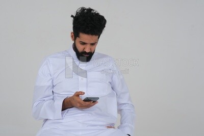 Saudi man in traditional Saudi dress, holding a mobile phone and checking it, Saudi model, white background
