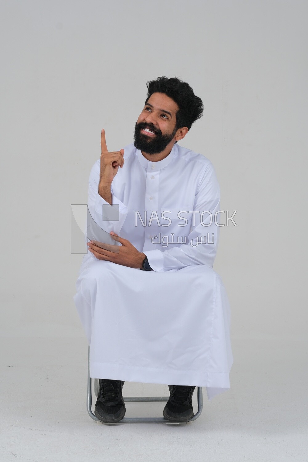 close-up of a Saudi man in traditional Saudi dress, sitting on a chair pointing up, Saudi model, white background