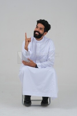 close-up of a Saudi man in traditional Saudi dress, sitting on a chair pointing up, Saudi model, white background