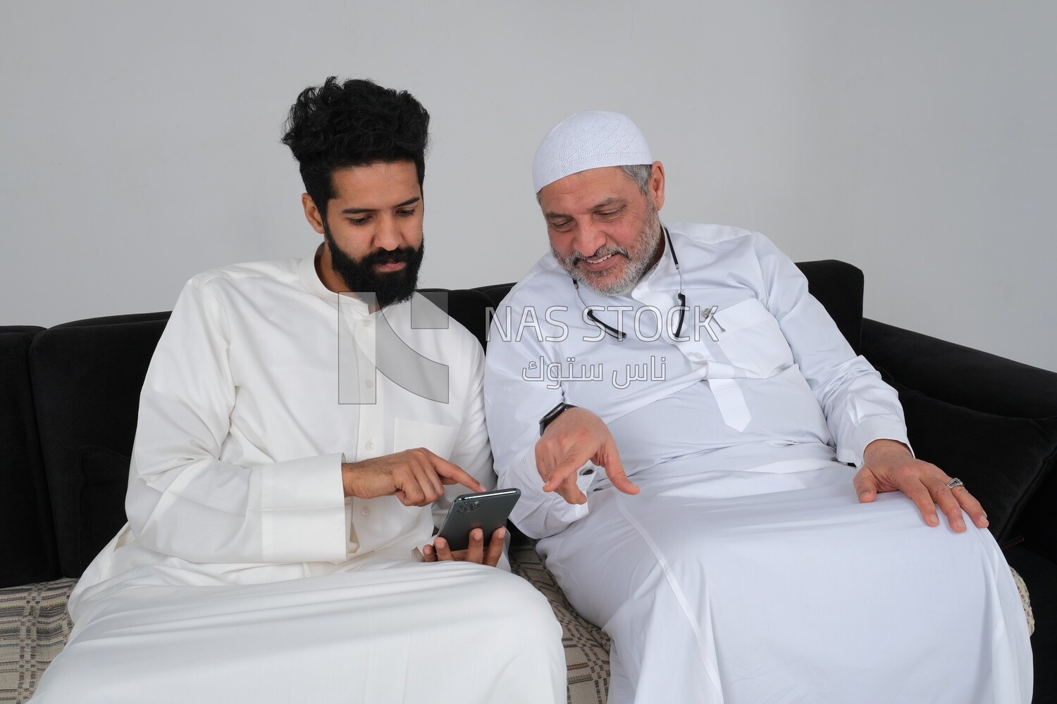 رجل سعودي يقضي الوقت مع والده جالسًا على الأريكة ، مشيرًا إلى الهاتف ، خلفية بيضاء ، موديل سعودى