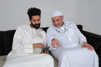Saudi man spending time with his father sitting on the sofa, pointing at the phone, white background, Saudi model