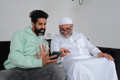 Saudi man spending time with his father sitting on the sofa, pointing at the phone, white background, Saudi model