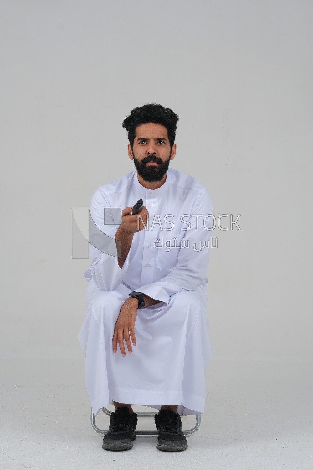 Saudi man wearing a Saudi dress, sitting on a chair, holding the remote control in his hand, spending good time, watching TV, happy facial gestures, spending fun times, entertainment, movies and cin