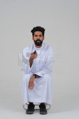 Saudi man wearing a Saudi dress, sitting on a chair, holding the remote control in his hand, spending good time, watching TV, happy facial gestures, spending fun times, entertainment, movies and cin