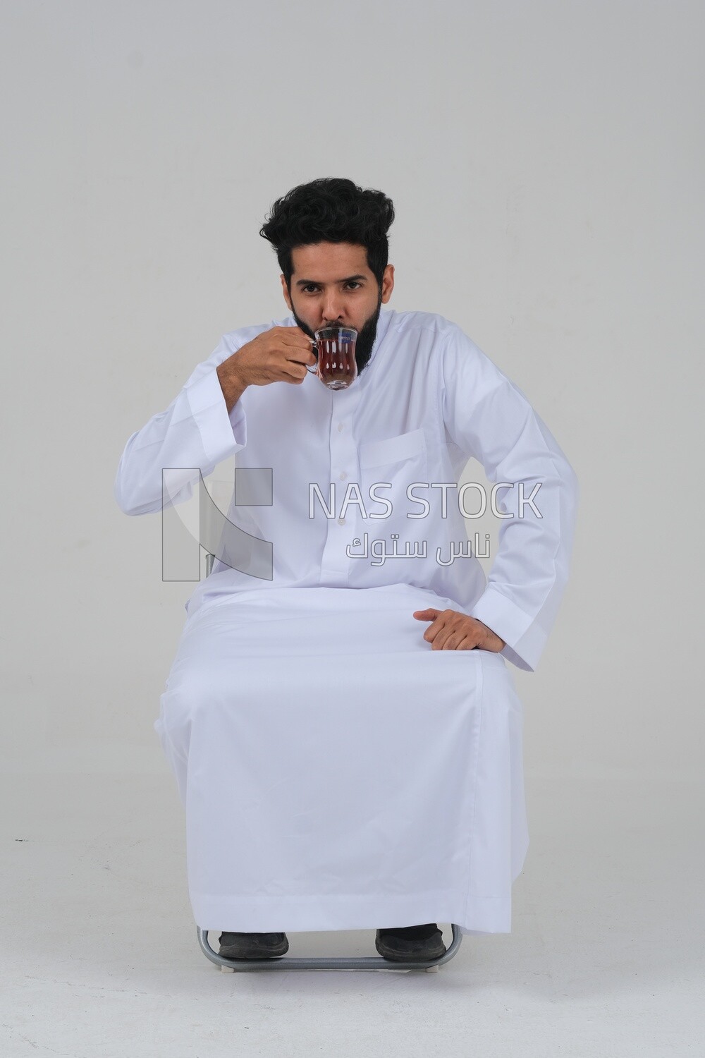 Saudi man wearing a white dress sitting on a chair, carrying a cup of tea and looking at it, self-confidence, white background, Saudi model
