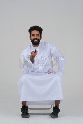 Saudi man wearing a Saudi dress, sitting on a chair, holding the remote control in his hand, spending good time, watching TV, happy facial gestures, spending fun times, entertainment, movies and cin
