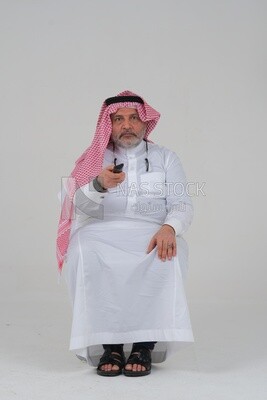 Saudi man wearing a Saudi dress, sitting on a chair, holding the remote control in his hand, spending good time, watching TV, happy facial gestures, spending fun times, entertainment, movies and cin