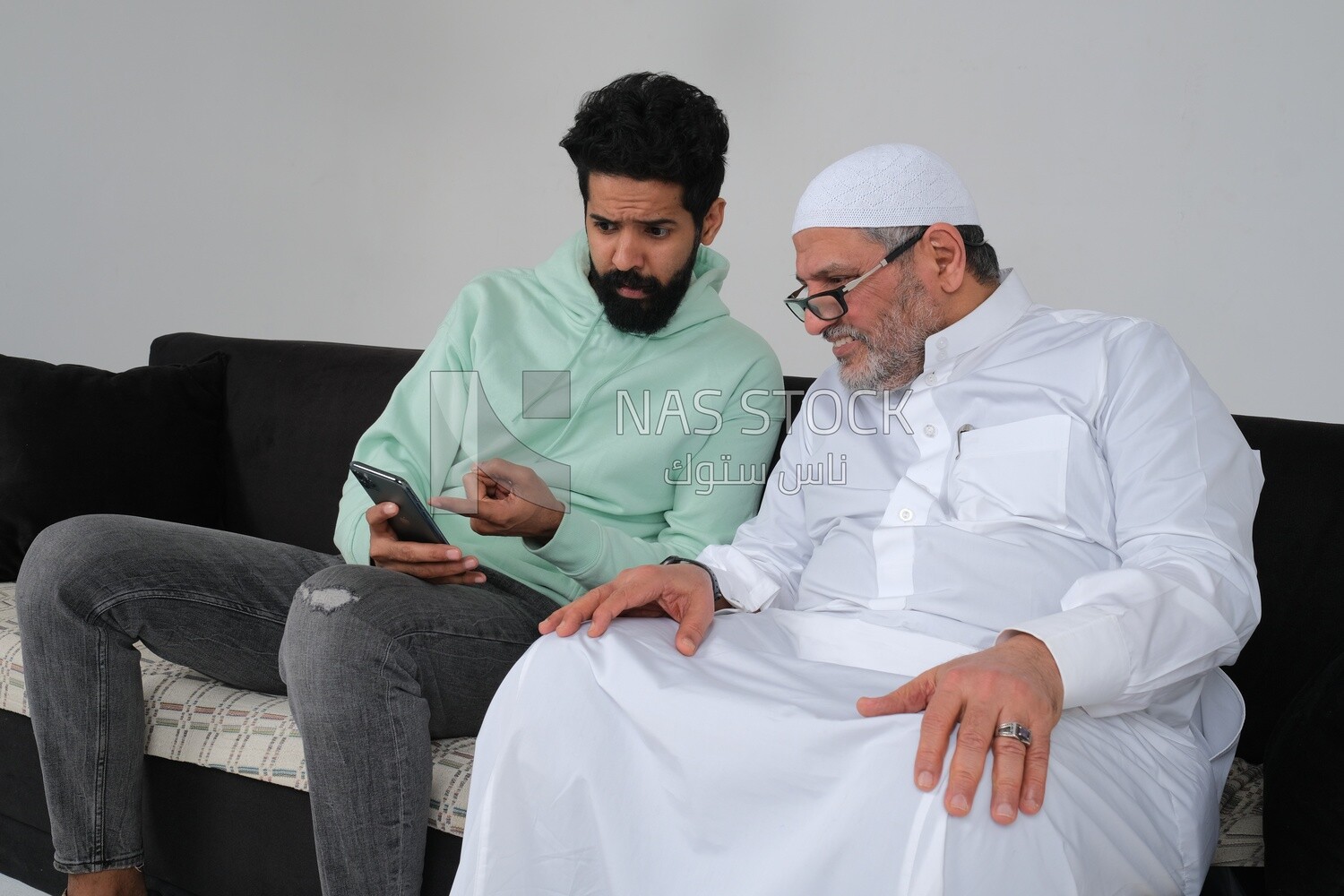 Saudi man spending time with his father sitting on the sofa, pointing at the phone, white background, Saudi model