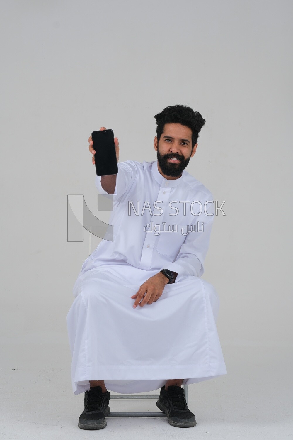 close-up of a Saudi man in traditional Saudi dress, using his mobile phone, sitting on a chair, Saudi model, white background