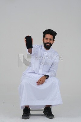 صورة مقربة لرجل سعودي يرتدي الزي السعودي التقليدي،يستخدم هاتفه المحمول ، يجلس على الكرسى ، موديل سعودى ، خلفية بيضاء