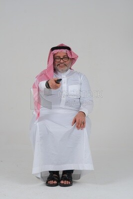 Saudi man wearing a Saudi dress, sitting on a chair, holding the remote control in his hand, spending good time, watching TV, happy facial gestures, spending fun times, entertainment, movies and cin
