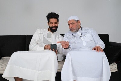 Saudi man spending time with his father sitting on the sofa, pointing at the phone, white background, Saudi model