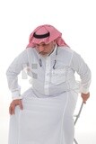 رجل سعودي مسن يحاول الجلوس على كرسي ، آلام الظهر والعمود الفقري ، إرهاق وإرهاق ، تشنجات عضلية ، مشاكل صحية ، خلفية بيضاء ، موديل سعودي.