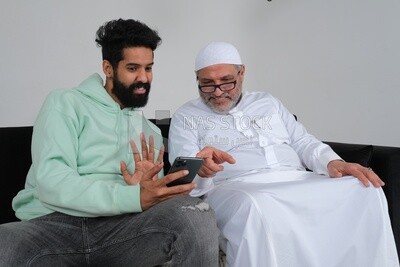 Saudi man spending time with his father sitting on the sofa, pointing at the phone, white background, Saudi model