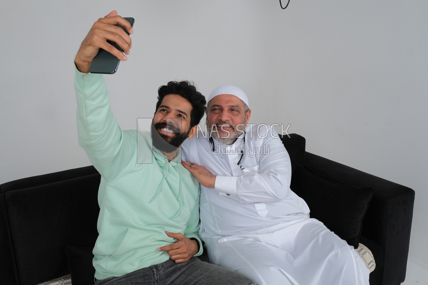 Saudi man spending time with his father sitting on the sofa, taking a selfie, white background, Saudi model