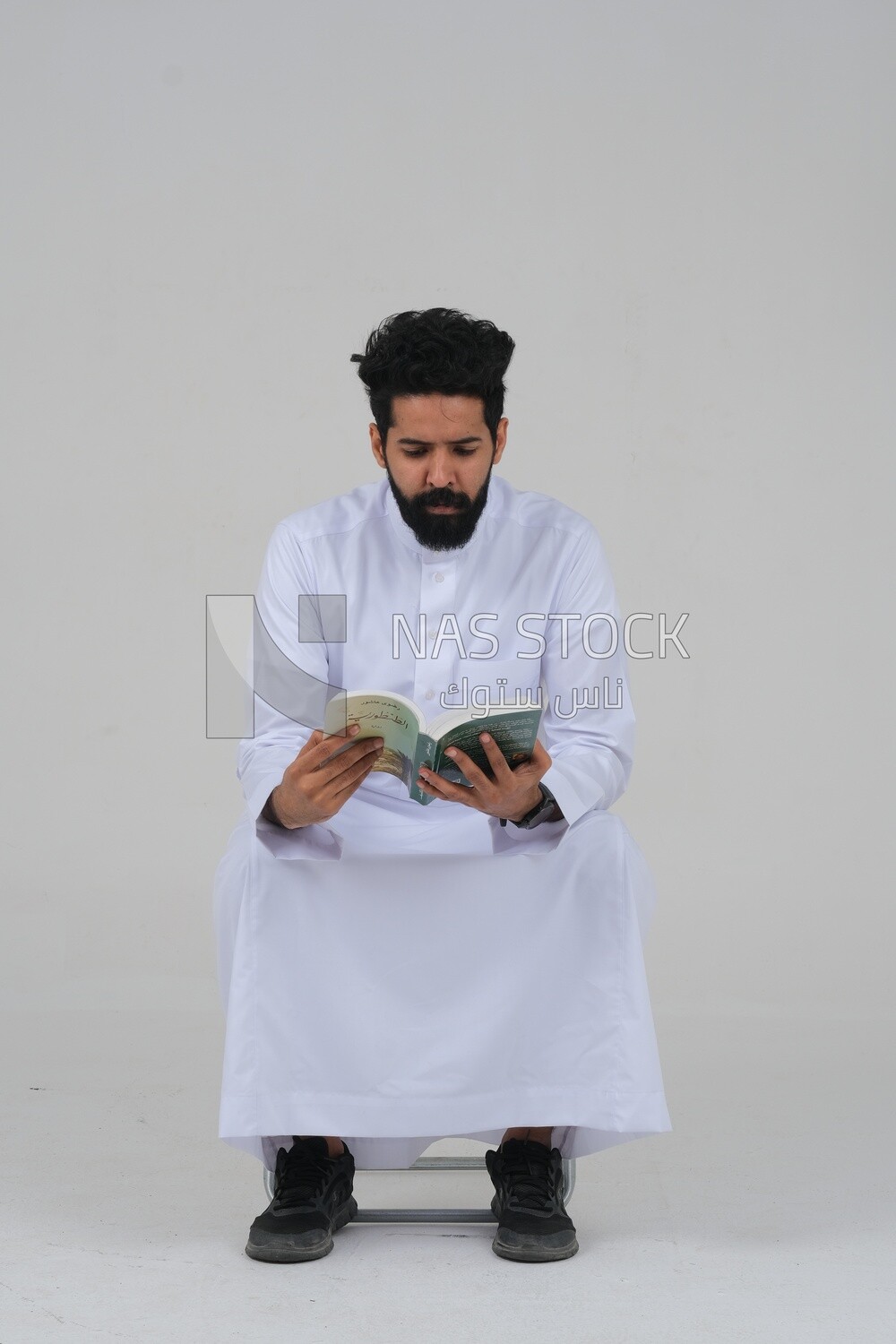 Saudi man wearing a white dress sitting on a chair, reading a book, self-confidence, white background, Saudi model