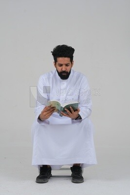 Saudi man wearing a white dress sitting on a chair, reading a book, self-confidence, white background, Saudi model