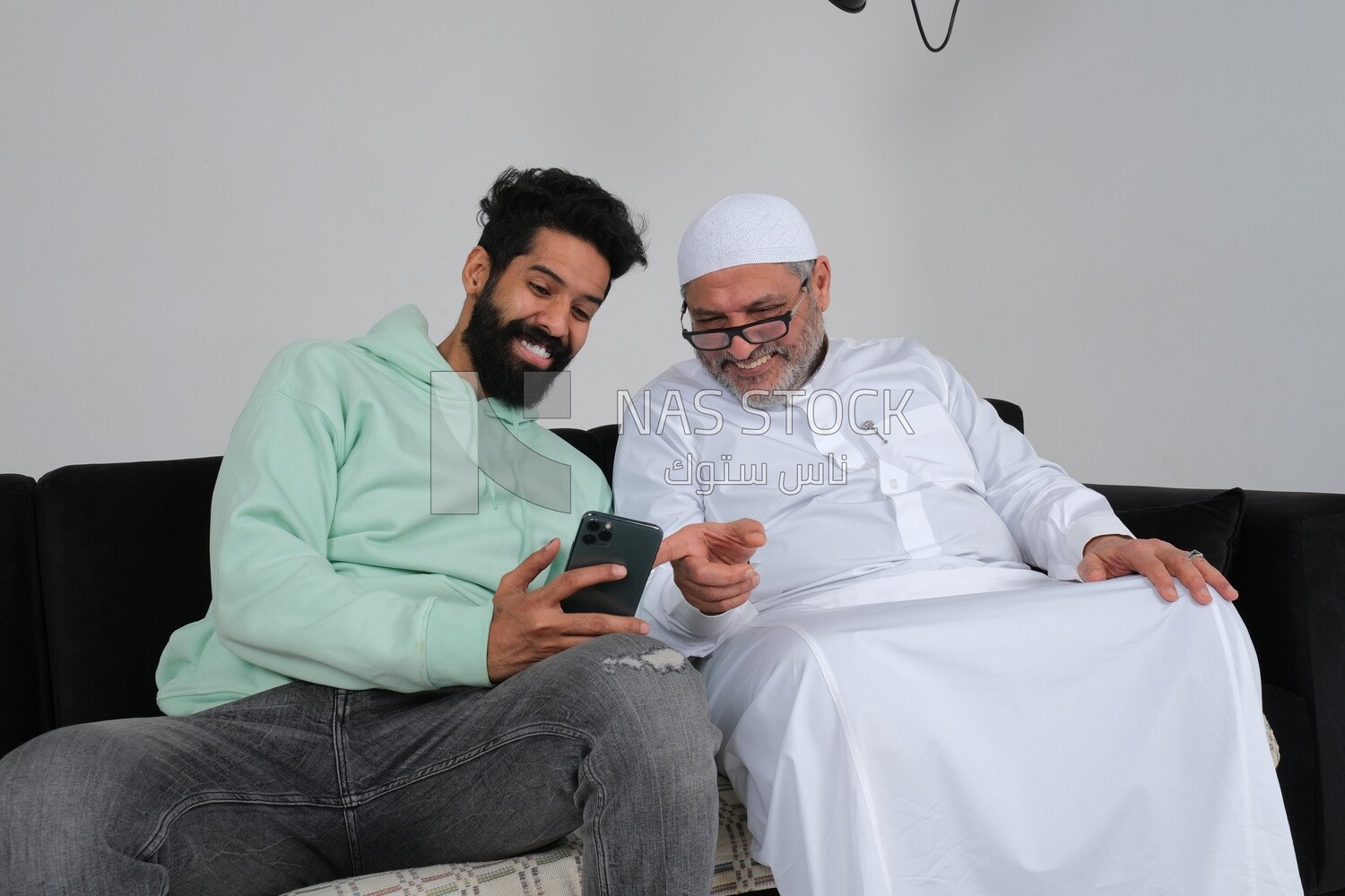 Saudi man spending time with his father sitting on the sofa, pointing at the phone, white background, Saudi model