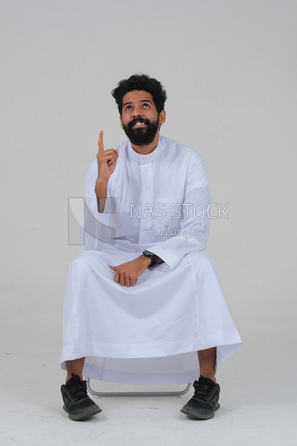 close-up of a Saudi man in traditional Saudi dress, sitting on a chair pointing up, Saudi model, white background