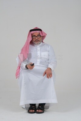 Saudi man wearing a Saudi dress, sitting on a chair, holding the remote control in his hand, spending good time, watching TV, happy facial gestures, spending fun times, entertainment