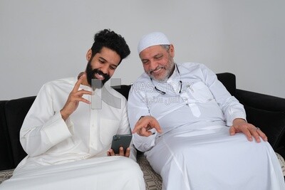Saudi man spending time with his father sitting on the sofa, pointing at the phone, white background, Saudi model