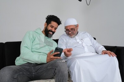 رجل سعودي يقضي الوقت مع والده جالسًا على الأريكة ، مشيرًا إلى الهاتف ، خلفية بيضاء ، موديل سعودى