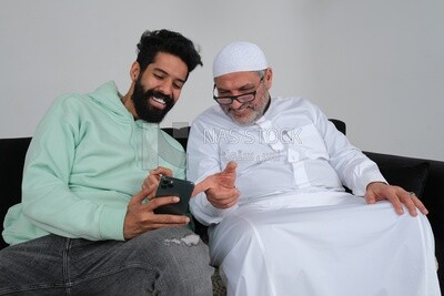 Saudi man spending time with his father sitting on the sofa, pointing at the phone, white background, Saudi model