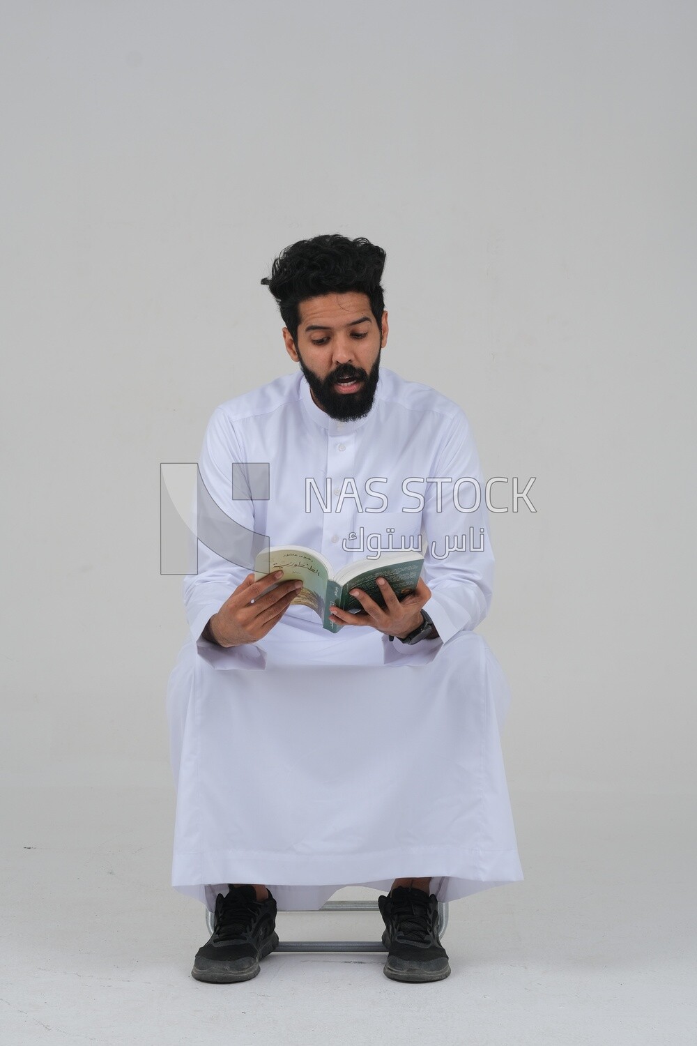 Saudi man wearing a white dress sitting on a chair, reading a book, self-confidence, white background, Saudi model