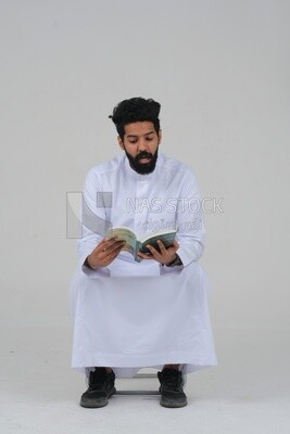 Saudi man wearing a white dress sitting on a chair, reading a book, self-confidence, white background, Saudi model