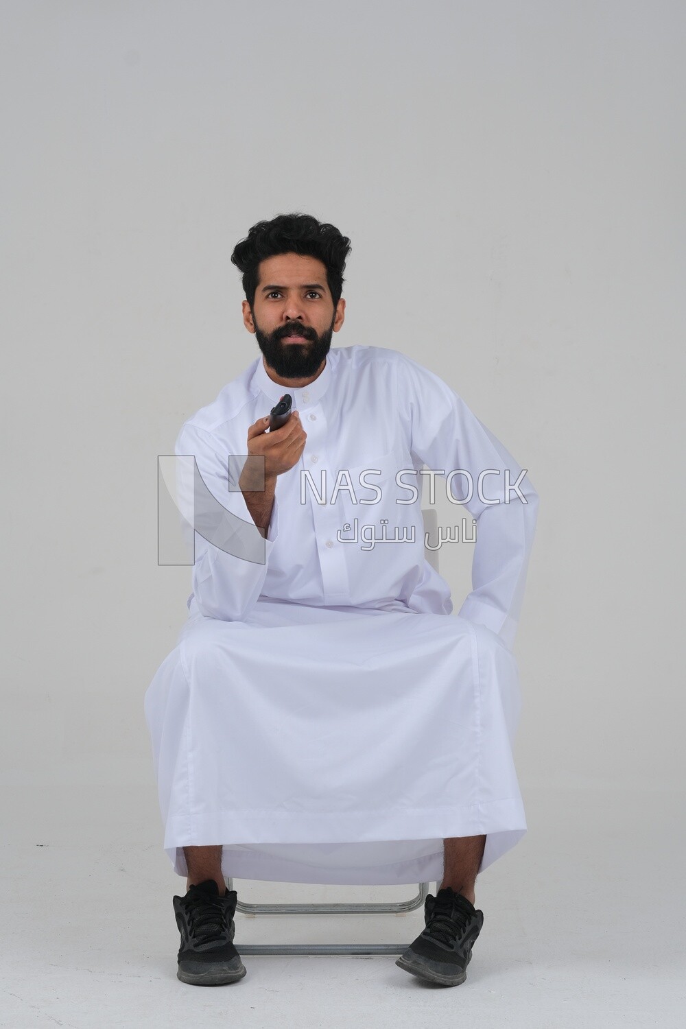 Saudi man wearing a Saudi dress, sitting on a chair, holding the remote control in his hand, spending good time, watching TV, happy facial gestures, spending fun times, entertainment